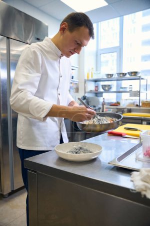 Foto de Empleado de cocina pone espaguetis negros en una salsa especial en un plato, trabaja en un uniforme cómodo. - Imagen libre de derechos