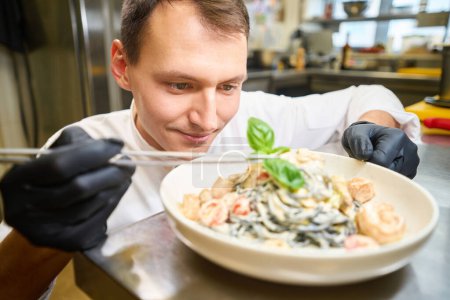 Foto de El hombre decora espaguetis en salsa con hojas de albahaca, trabaja con guantes protectores - Imagen libre de derechos