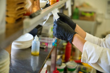 Téléchargez les photos : Travailleur de cuisine en gants de protection noirs regarde à travers un reçu, il y a de la vaisselle et de la nourriture sur les étagères - en image libre de droit