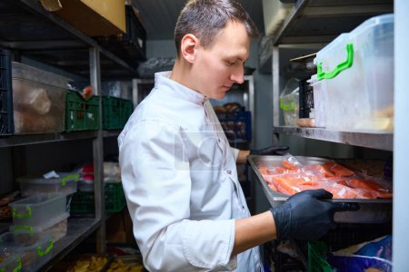 Foto de Empleado de cocina con una bandeja de pescado rojo en las manos en un refrigerador restaurante, alrededor de estantes con comida - Imagen libre de derechos