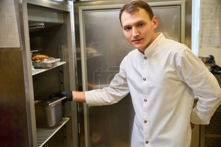 Foto de Cocinar en uniforme pone un recipiente de alimentos en el refrigerador, hay muchos recipientes con alimentos en los estantes - Imagen libre de derechos