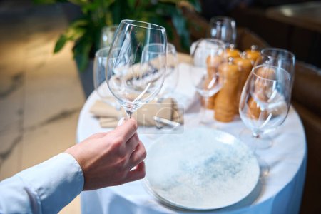 Waiter holds a large glass in his hands, he is setting a table in the restaurant hall