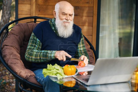 Rentner bereitet auf der Veranda vor dem Laptop einen Salat zu, er sitzt in einem gemütlichen Stuhl
