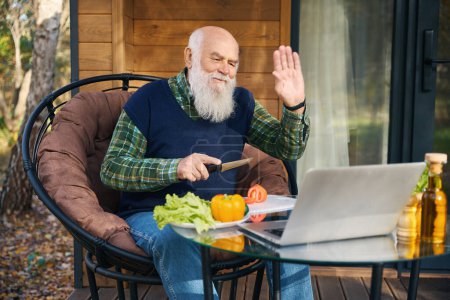 Alter Mann bereitet Salat auf der Veranda zu und kommuniziert online, er sitzt in einem gemütlichen Stuhl