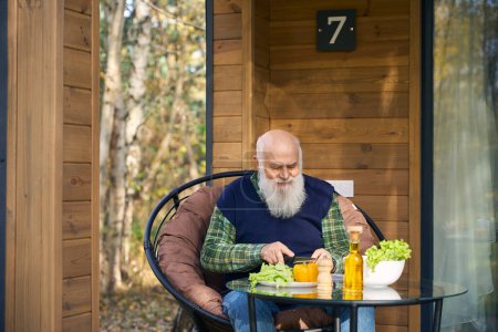 Alter Mann bereitet Gemüsesalat auf der Veranda zu, er sitzt in einem gemütlichen Stuhl