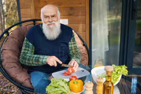 Old man is chopping vegetables for a salad, he is sitting on a cozy veranda in a garden chair