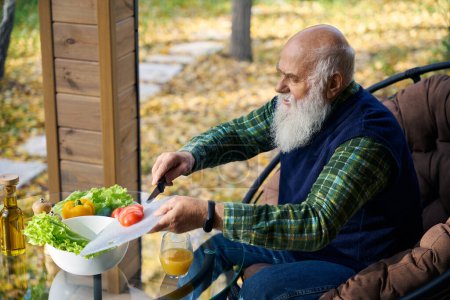 Älterer Mann hackt Gemüse für einen Salat, er sitzt auf einer gemütlichen Veranda im Gartenstuhl