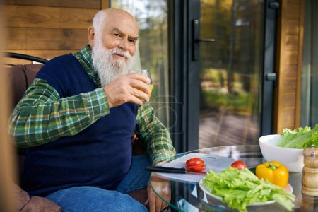 Foto de Viejo sonriente está preparando una ensalada de verduras, él está sentado en una acogedora terraza con un vaso de jugo - Imagen libre de derechos