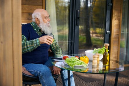 Bärtiger alter Mann bereitet Gemüsesalat zu, er sitzt auf einer gemütlichen Veranda mit einem Glas Saft