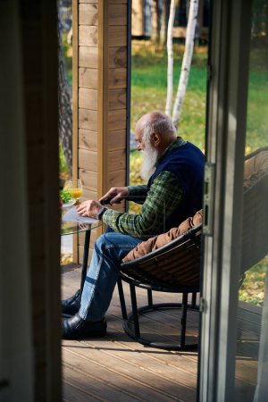 Graubärtiger alter Mann bereitet Gemüsesalat zu, er sitzt auf einer gemütlichen Veranda im Gartenstuhl