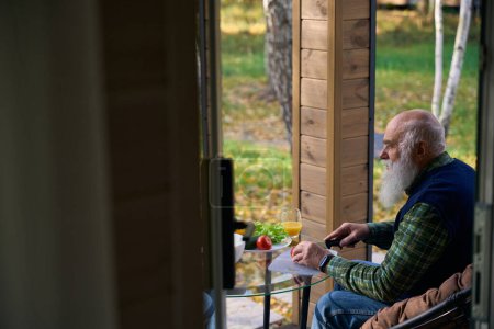 Grauhaariger alter Mann bereitet Gemüsesalat zu, er sitzt auf einer gemütlichen Veranda im Gartenstuhl