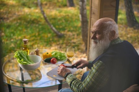 Grauhaariger alter Mann hackt Gemüse für einen Salat, er sitzt auf einer gemütlichen Veranda im Gartenstuhl