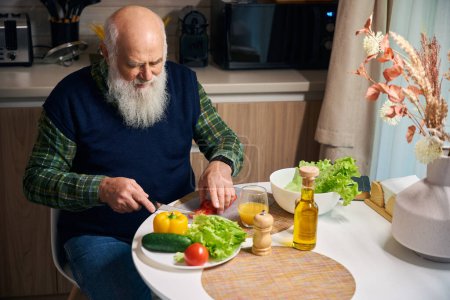 Téléchargez les photos : Vieil homme à la table de cuisine coupe des légumes pour une salade, il est dans des vêtements de maison confortables - en image libre de droit
