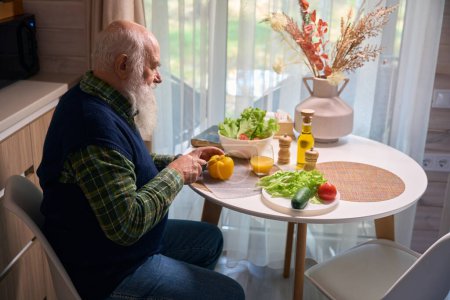 Foto de Hombre de barba gris en la mesa de la cocina corta verduras para una ensalada, lleva un chaleco azul cálido - Imagen libre de derechos