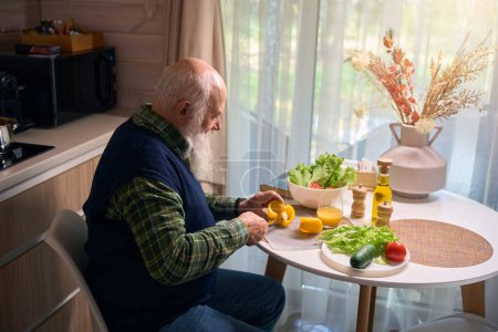 Téléchargez les photos : Homme âgé à la table de la cuisine coupe des légumes pour une salade, il porte un gilet bleu chaud - en image libre de droit