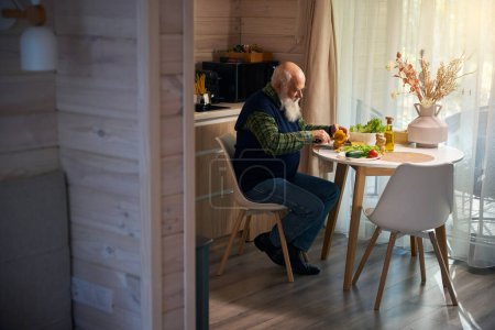 Foto de El viejo en la mesa de la cocina está cortando verduras para una ensalada, lleva un chaleco azul cálido. - Imagen libre de derechos