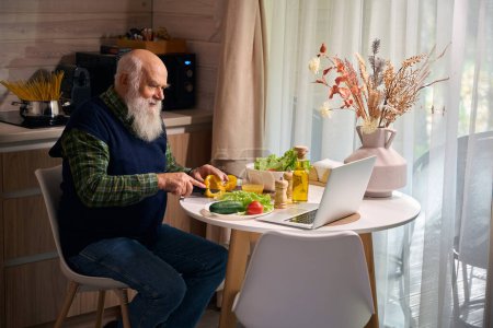 Foto de Viejo está preparando una ensalada y charlando en línea, él está usando un chaleco azul cálido - Imagen libre de derechos