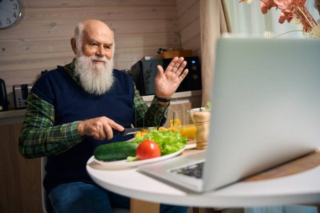 Foto de Viejo preparando una ensalada mientras charla en línea en casa, él está usando un chaleco azul cálido - Imagen libre de derechos