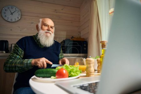 Foto de Anciano está preparando una ensalada y la comunicación en línea, hay un ordenador portátil moderno en la mesa - Imagen libre de derechos