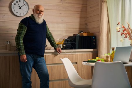 Elderly man in a warm vest stands in the kitchen, on the kitchen table there is a modern laptop