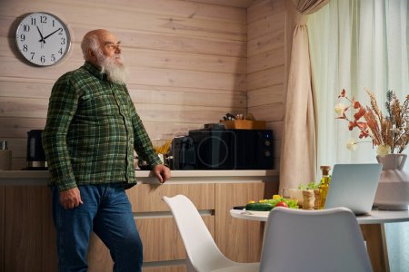 Photo for Old man in a warm checkered shirt stands in the kitchen, on the kitchen table there is a modern laptop - Royalty Free Image