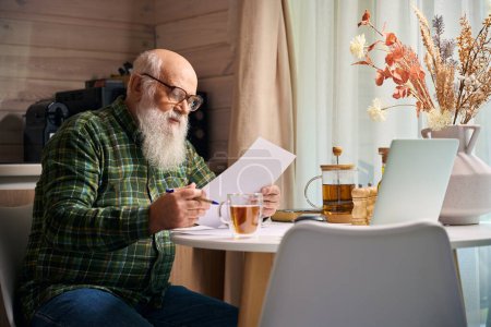 Photo for Grandfather sitting down at the kitchen table with laptop and documents, on table there was a composition of dried flowers - Royalty Free Image