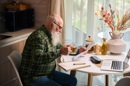 Téléchargez les photos : Homme âgé travaille à la table de cuisine sur un ordinateur portable, il y a une composition de fleurs séchées sur la table - en image libre de droit