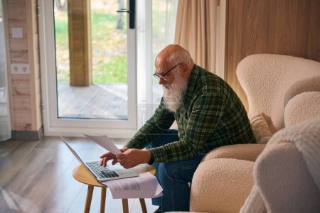 Anciano trabaja con documentos en un portátil, se sienta en una silla cerca de la ventana francesa