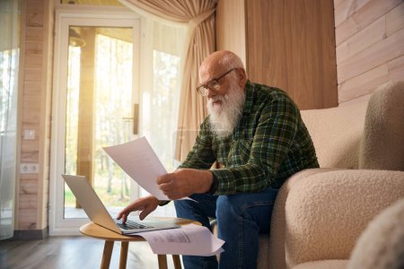 Foto de Viejo canoso trabaja con documentos en un portátil, se sienta en una silla cerca de la ventana francesa - Imagen libre de derechos