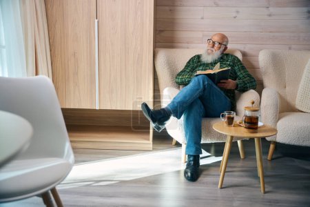 Téléchargez les photos : Pensionné s'assoit avec un livre dans une chaise confortable, la chambre a un design minimaliste - en image libre de droit