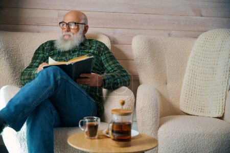 Téléchargez les photos : Vieil homme avec des lunettes installé avec un livre dans une chaise confortable, la chambre a un design minimaliste - en image libre de droit