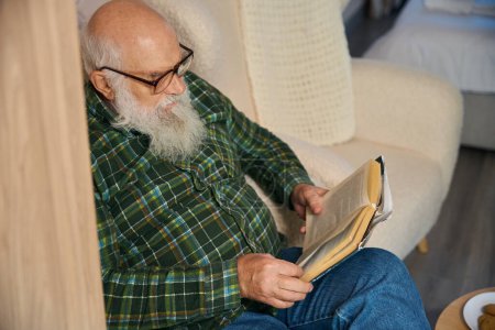 Téléchargez les photos : Vieil homme dans une chemise à carreaux chaud lit un livre, il est confortablement assis dans une zone de loisirs - en image libre de droit