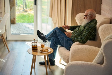 Anciano con gafas se sienta con un libro en una silla cómoda, la ventana francesa da a la terraza