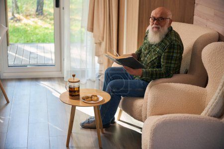 Téléchargez les photos : Vieil homme à barbe grise à lunettes installé avec un livre près de la porte-fenêtre, il s'assoit dans une chaise confortable - en image libre de droit