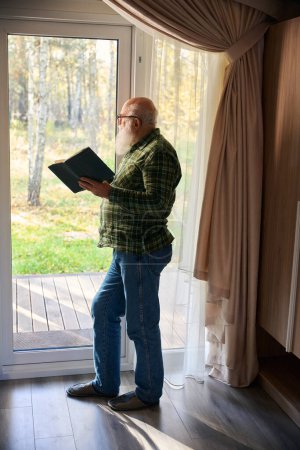 Téléchargez les photos : Vieil homme debout avec un livre à la porte-fenêtre, il admire la nature - en image libre de droit