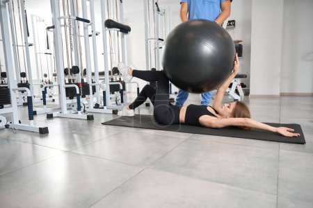 Photo for Woman performs exercises with a fitball under the supervision of a trainer - Royalty Free Image