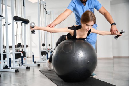 Foto de Paciente en una clínica de kinesioterapia realiza ejercicios con un fitball y pesas bajo la supervisión de un instructor - Imagen libre de derechos