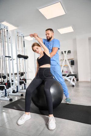 Photo for Woman performs muscle stretching exercises with a fitball under the supervision of an instructor - Royalty Free Image
