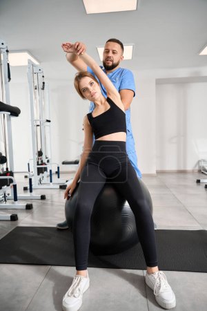 Photo for Instructor helps a patient perform exercises on a large fitball, a woman in a training suit - Royalty Free Image