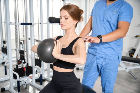 Foto de Instructor ayuda a un paciente a realizar ejercicios con una pelota, una mujer en un traje de entrenamiento - Imagen libre de derechos
