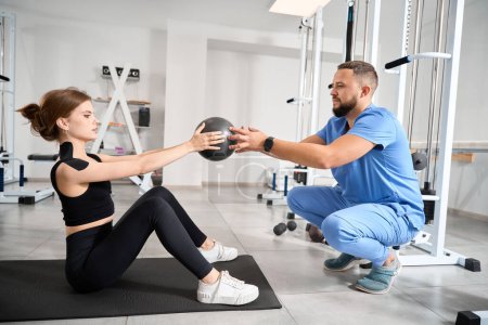 Photo for Woman performs exercises with a ball under the supervision of a kinesiologist, a man in a blue uniform - Royalty Free Image
