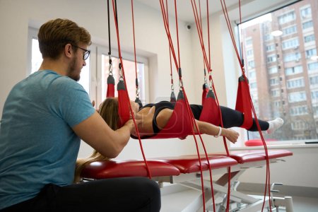 Male rehabilitologist fixes a woman in a suspension loop system, using a modern healing technique