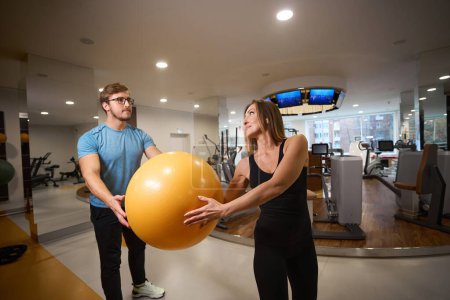Photo for Female and male are holding a yellow fitball in their hands, they are in a modern gym - Royalty Free Image
