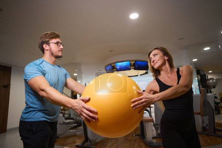 Photo for Man and a woman are holding a yellow fitball in their hands, they are in a modern gym - Royalty Free Image