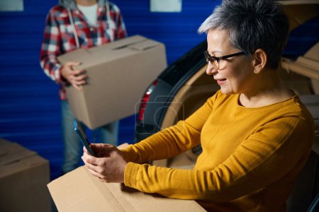 Photo for Woman looks at her phone holding box on her lap while a man takes box out of their trunk - Royalty Free Image
