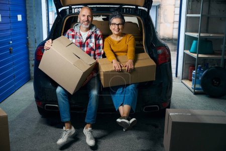 Photo for Man and a woman with short hair are holding cardboard boxes and sitting near an open trunk - Royalty Free Image