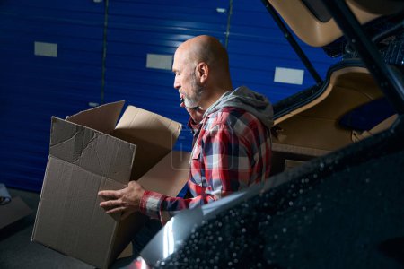 Photo for Adult guy in a shirt takes out a cardboard box with things from the trunk of a car. - Royalty Free Image