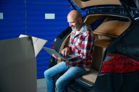 Foto de El tipo con una camisa a cuadros está escribiendo en un portátil mientras está sentado en el maletero abierto de un coche. - Imagen libre de derechos
