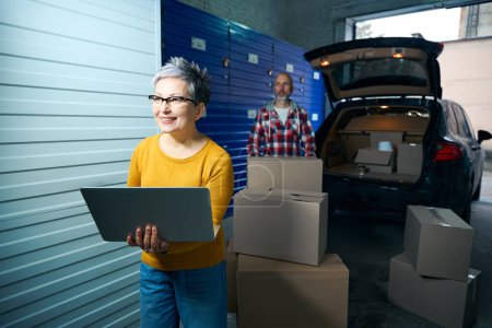 Photo for Woman with a short hair looks at a laptop while a man pulls boxes out of a car - Royalty Free Image