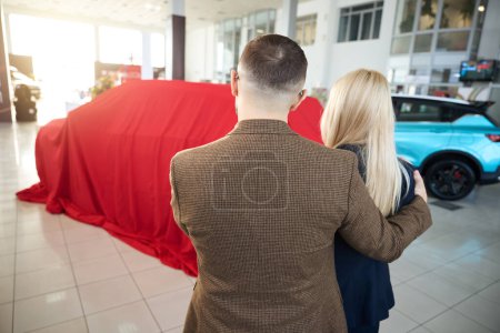 Foto de Esposo y esposa miran un coche nuevo que está envuelto en una caja de regalo roja - Imagen libre de derechos
